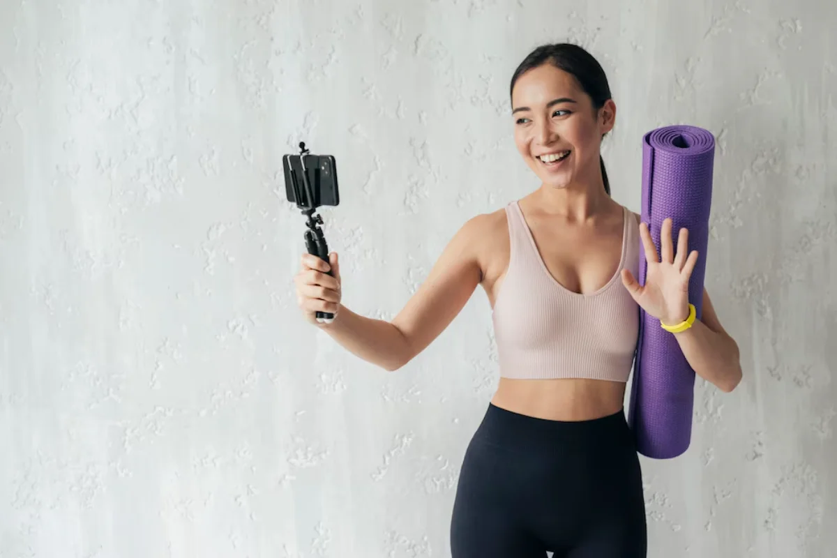 Young woman holding a yoga mat and recording a video with a smartphone on a selfie stick, smiling and waving.
