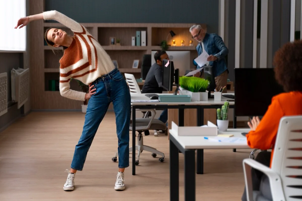 A woman stretches in a bustling office environment, colleagues working around her.