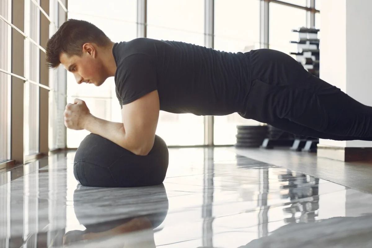 Man doing a plank exercise on a glossy floor in a bright, spacious gym.