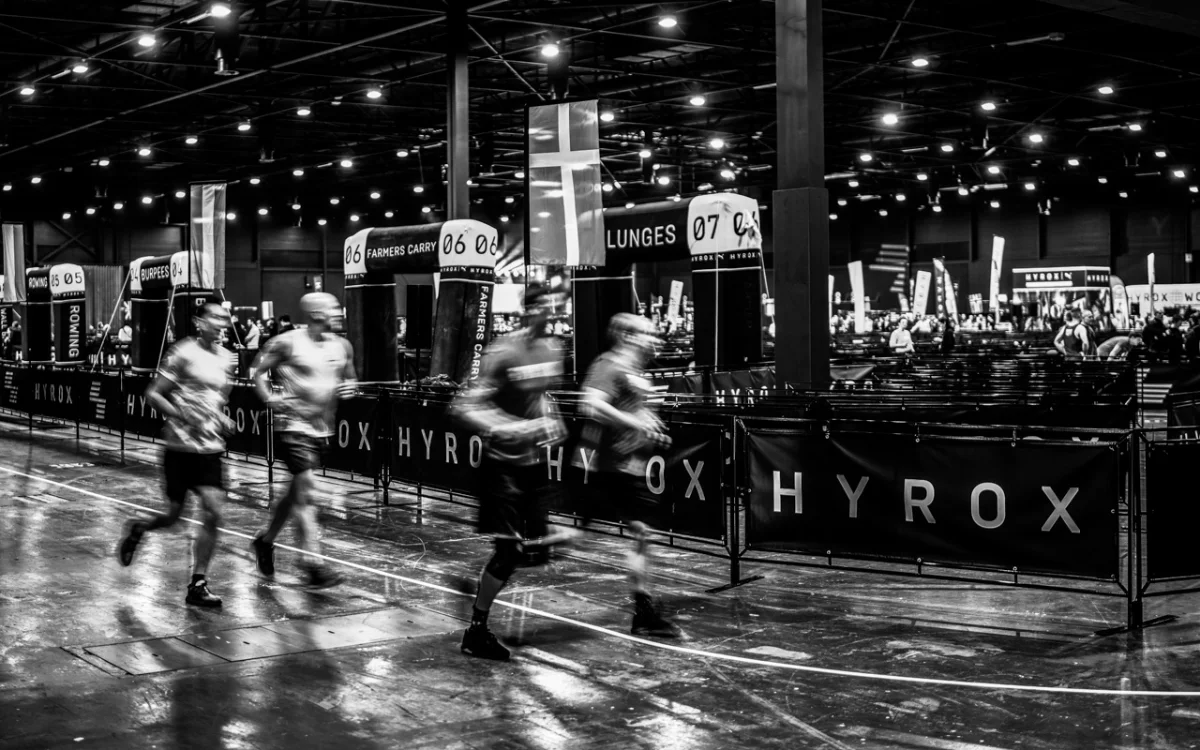 Blurred motion of athletes racing in a large, indoor fitness competition with banners and station markers.