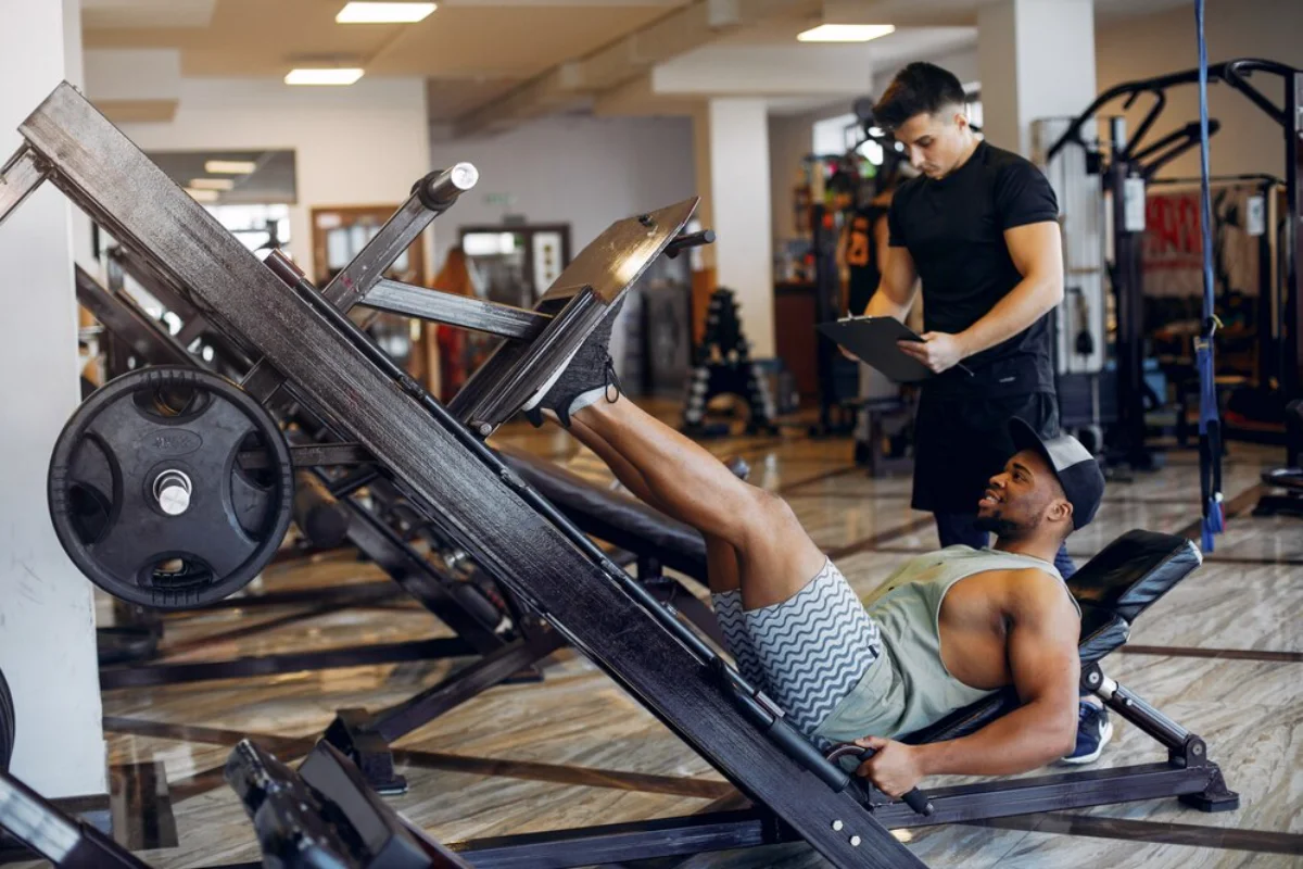 Man performing leg presses at a gym while a trainer observes and takes notes.