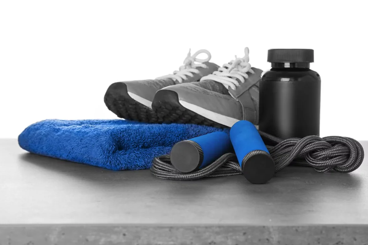 Workout essentials including sneakers, a blue towel, a black protein shaker, and a skipping rope on a grey surface.