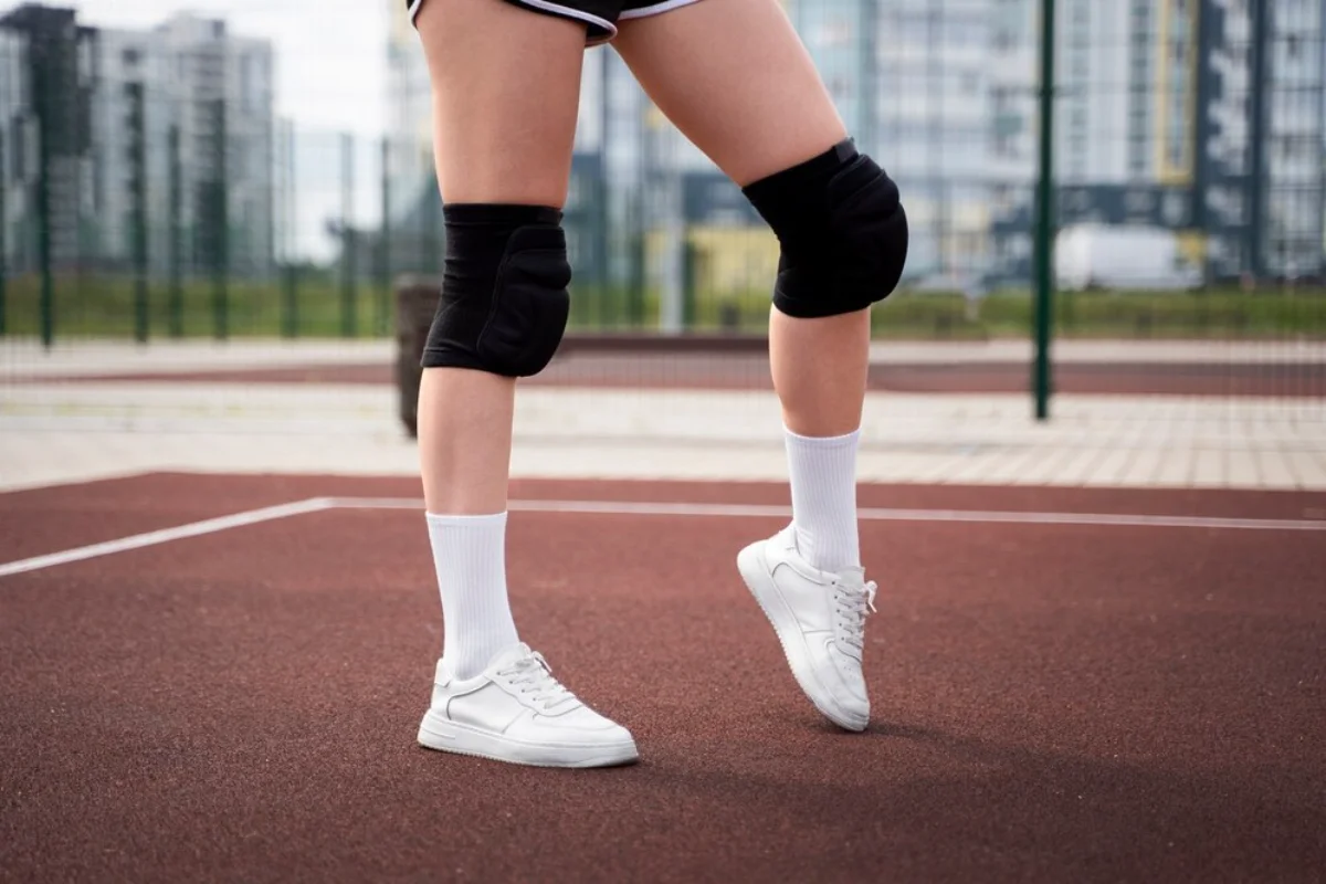 Close-up of a person's legs wearing knee pads and white sneakers on a sports court, urban buildings in the background.