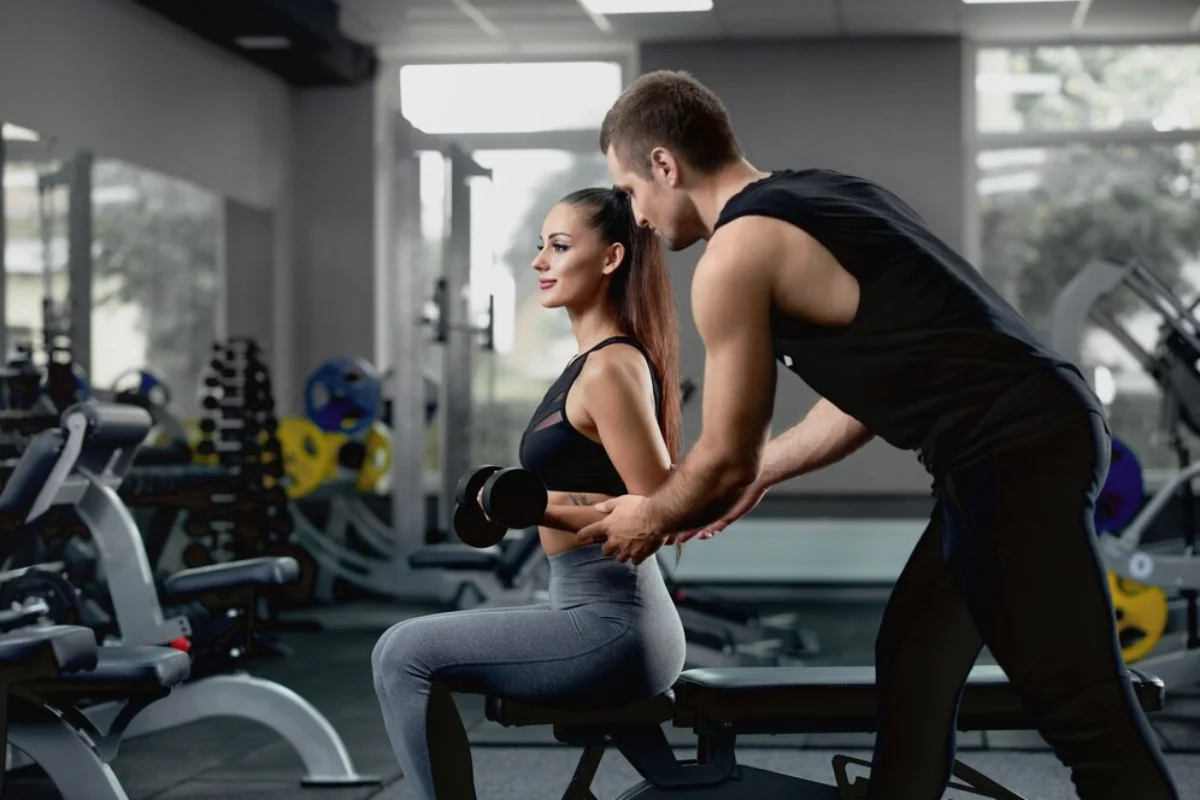 Personal trainer assisting a woman with dumbbell exercises in a gym.