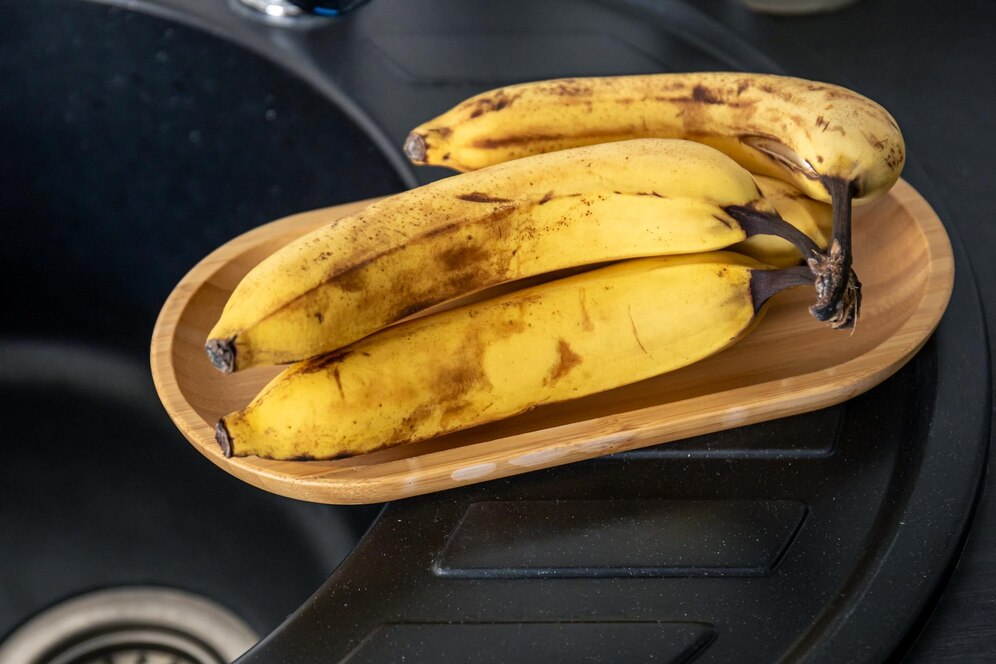 A wooden bowl with ripe, spotty bananas on a black kitchen countertop.