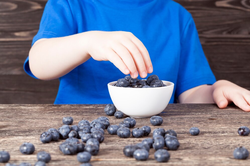 Child in a blue shirt picking blueberries from a white bowl on a wooden table.