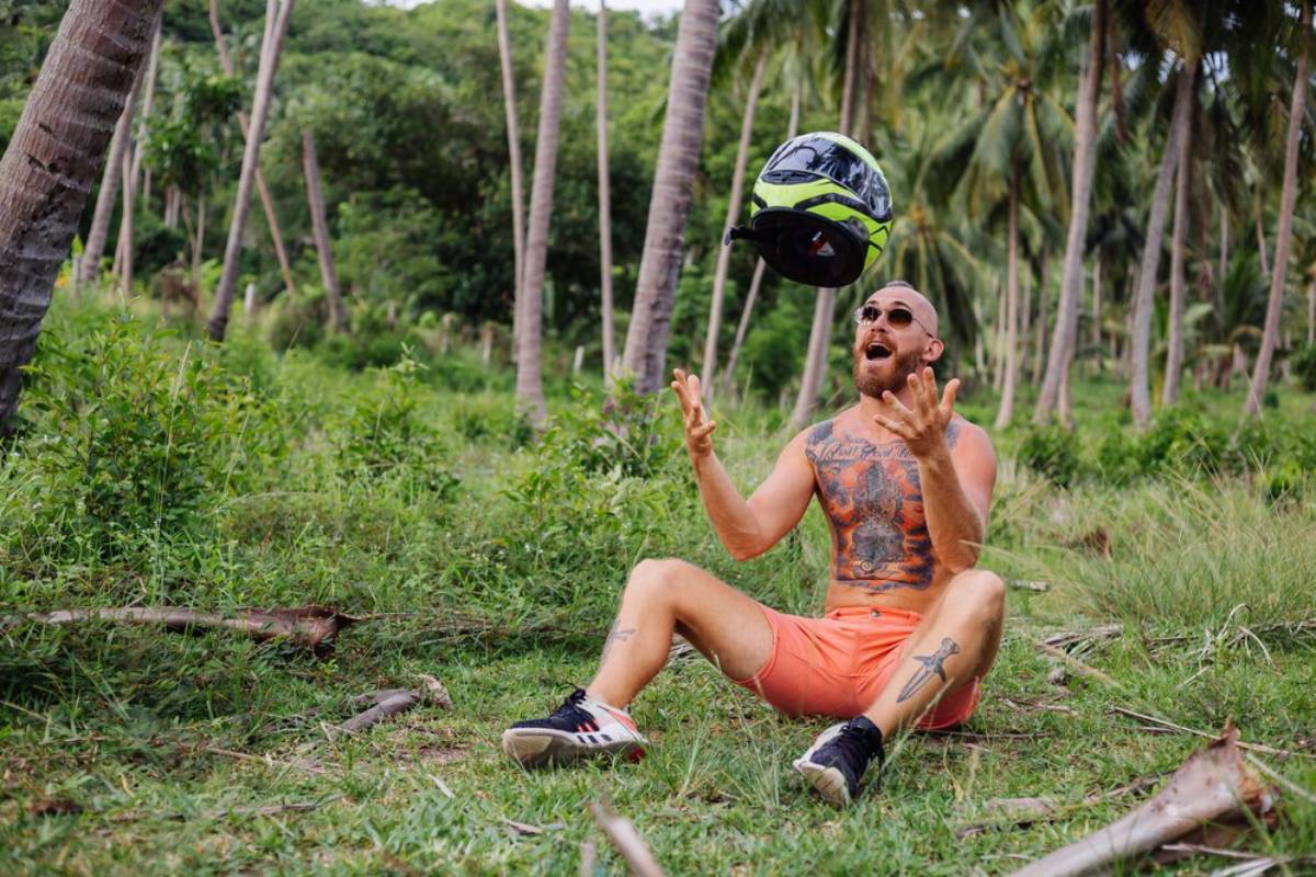 Man in orange shorts and tank top tossing a motorcycle helmet in the air, surrounded by palm trees.