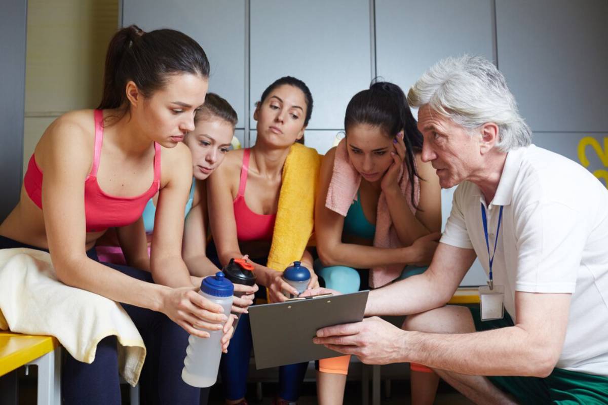 A senior male coach showing a tablet to four focused young female athletes in a gym setting.