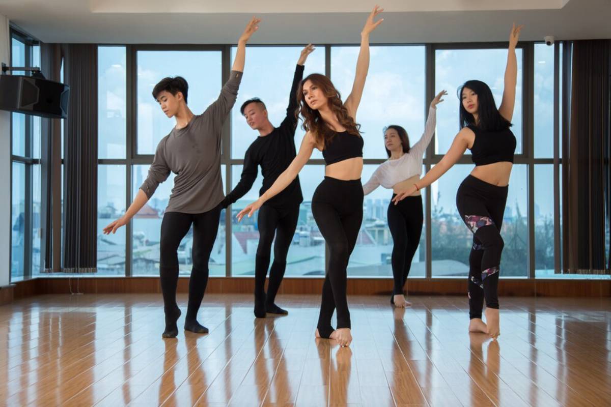 Five adults performing a dance routine in a bright studio with large windows.