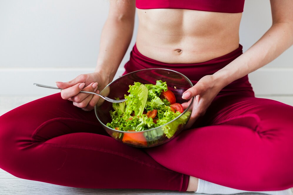 A woman in red athletic wear sits cross-legged, holding a bowl of fresh salad with lettuce and tomatoes, ready to eat.