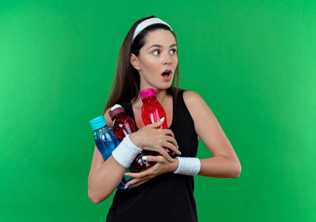 Surprised woman in sportswear holding multiple colorful water bottles against a green background.