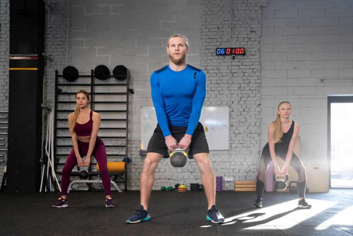 Two women and a man performing kettlebell exercises in a sunlit gym with white brick walls.