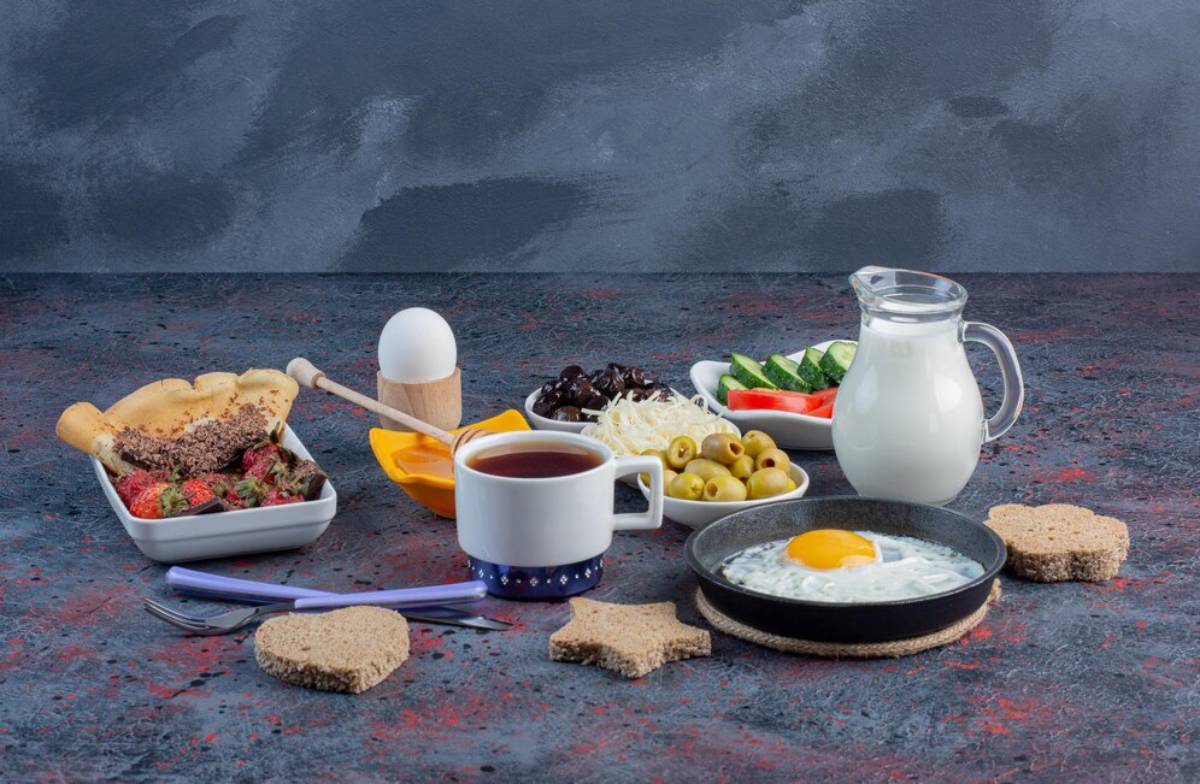 Assorted breakfast foods including egg, bread, olives, cheese, and a jug of milk on a textured table.