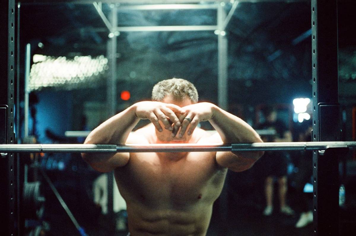 Man resting at the gym, holding his head while leaning on a barbell in a squat rack.