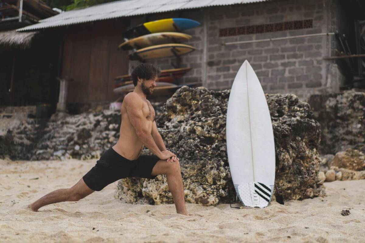 Man stretching on a beach next to a surfboard, with a surf shack and assorted surfboards in the background.