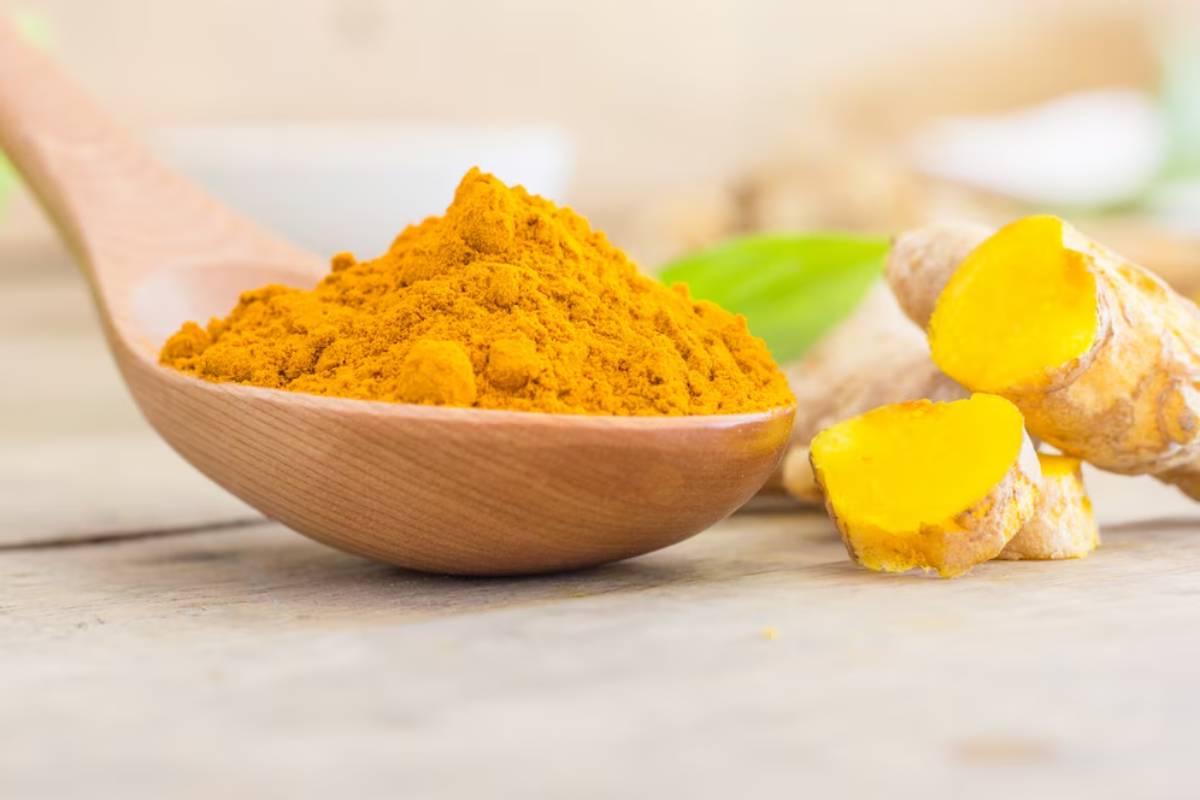 A wooden spoon holds bright orange turmeric powder, with fresh turmeric root pieces nearby, set on a rustic wooden surface.