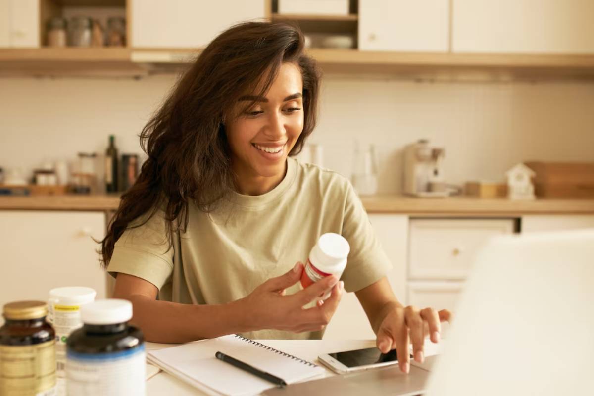 A woman smiling while holding a medication bottle and using a laptop in a kitchen setting.