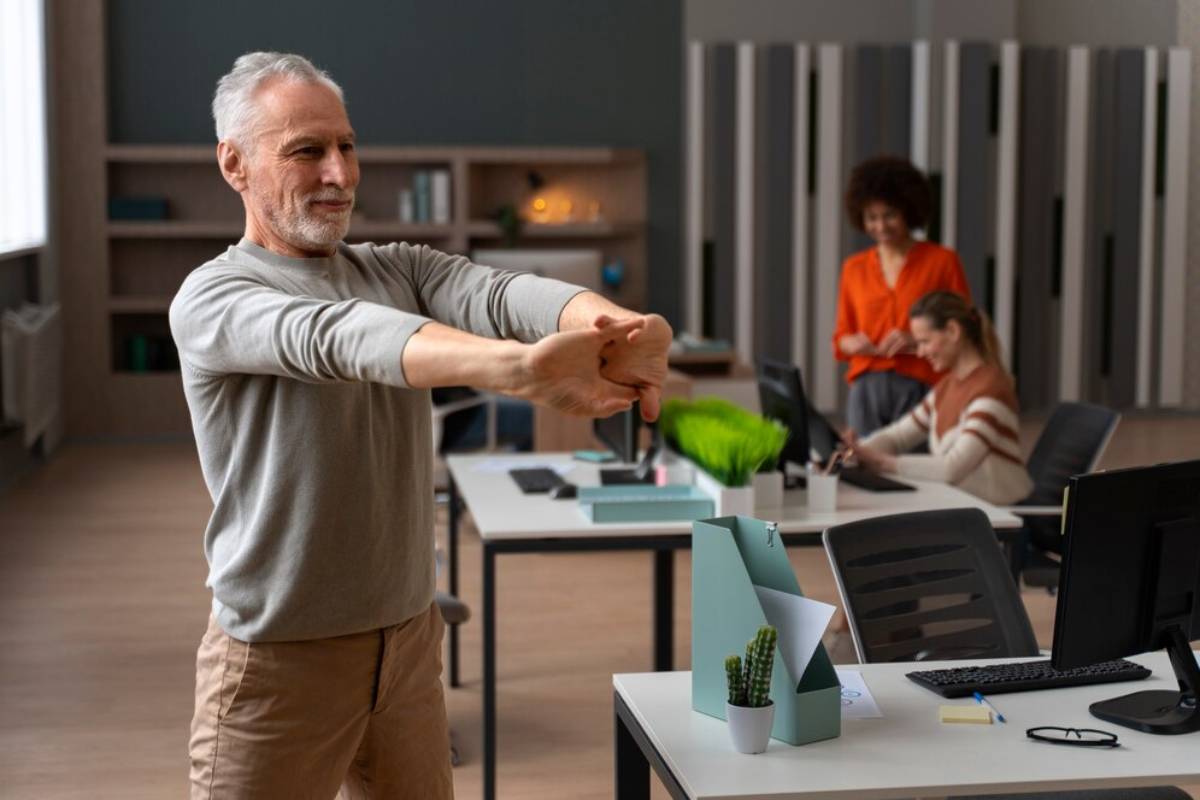 Senior man stretching his arms in an office environment with two female colleagues working in the background.
