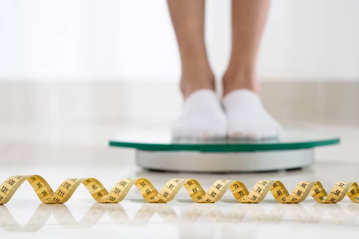 Person standing on a bathroom scale with a measuring tape in the foreground.