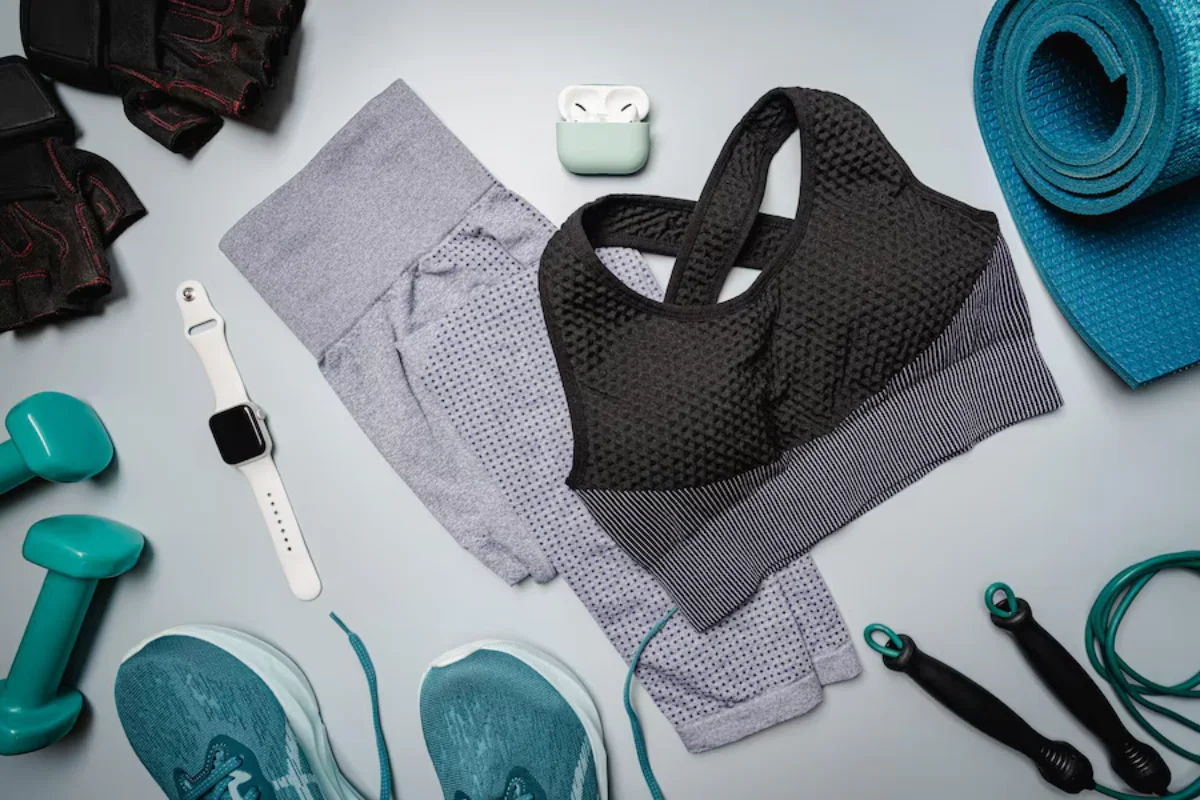 Assorted fitness gear including shoes, dumbbells, yoga mat, smartwatch, and clothing on a grey background.
